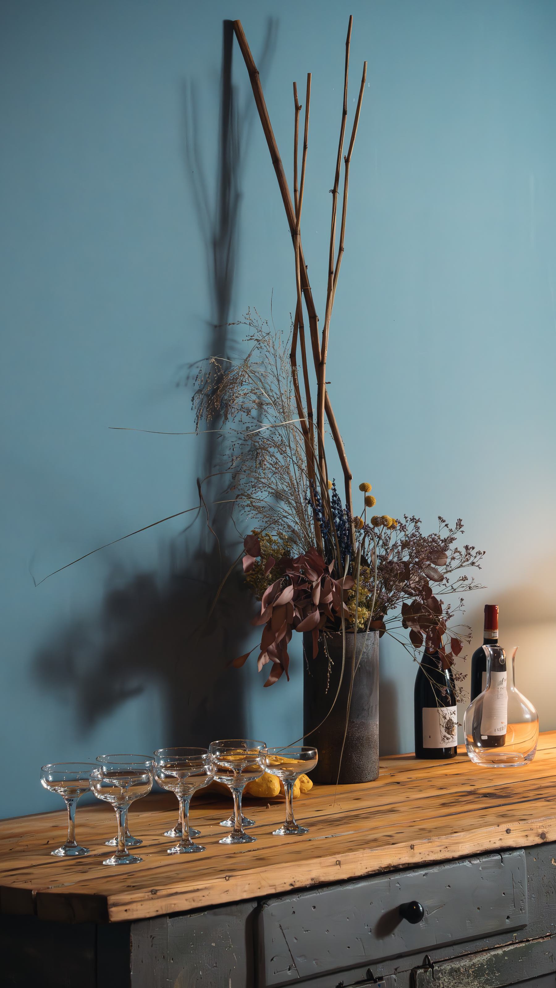 Coupégläser, Flaschen und Trockenblumen auf einer Holzplatte vor blauer Wand